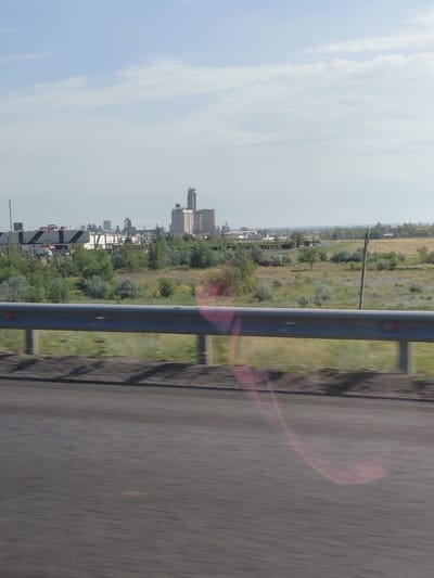The image shows an industrial landscape with a large, tall concrete structure in the distance, possibly a silo or a processing plant. In the foreground, a metal guardrail runs horizontally across the frame, with a road visible below it, showing motion blur. The background features a hazy sky with scattered clouds, and more buildings and greenery extending into the distance. The overall impression is one of a somewhat industrial or rural setting, possibly on the outskirts of a city. The presence of the guardrail and the blurred road suggest the photo was taken from a moving vehicle, likely on a highway or main road.