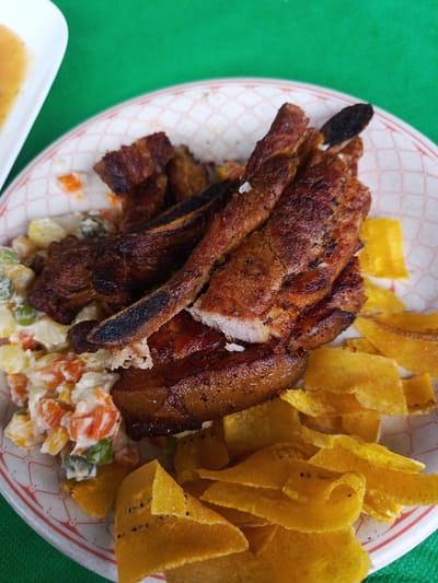 The image shows a plate of food, likely taken in Quevedo, Ecuador, based on the typical dish composition. The meal includes:

*   Grilled pork chops, some with visible ribs, displaying a dark char.
*   Tostones (fried plantain slices) arranged on the side.
*   A scoop of vegetable salad with visible peas, carrots, and possibly potato, mixed with a creamy dressing.

The food is served on a disposable paper plate with a checkered pattern. The background is a solid green color, possibly a tablecloth or outdoor setting. Part of another white plate can be seen in the upper left corner. No people are visible in the image. The overall lighting suggests a daytime shot, possibly outdoors.