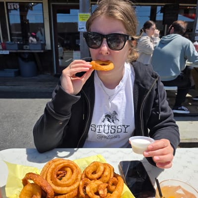 In Cannon Beach, Oregon, United States of America, a young woman with blonde hair and sunglasses is captured mid-bite, holding a golden-brown onion ring in her hand as she eats it. She wears a black zip-up hoodie over a white t-shirt that features a graphic of a lion and the word "STUSSY Sportswear". Her right hand, adorned with a ring on her index finger, holds the onion ring, while her left hand is visible near the onion rings on a yellow-lined tray in front of her. Beside the tray, a clear glass containing an amber-colored beverage with ice and a black straw sits next to a dark-colored smartphone. The scene unfolds outdoors at a table with a white, patterned surface. In the background, a casual dining establishment with large windows and signage is visible, with other patrons seated. The overall atmosphere suggests a relaxed meal in a sunny outdoor setting.