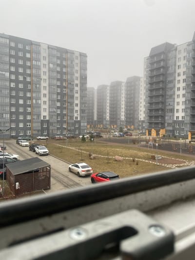 The image depicts an exterior view of a modern residential complex in Новогорелово, Russia, under heavily overcast or foggy conditions. Multiple multi-story apartment buildings, primarily gray and white with dark roof accents, line the background, with distant structures significantly obscured by the atmospheric haze. In the mid-ground, a paved road winds through a landscaped courtyard featuring sparse brown grass and small trees. Numerous vehicles are parked along the road and in designated spots; a white sedan and a red crossover with a black roof are prominent on the right, while a white SUV is visible on the left. A wooden slatted enclosure with a dark, sloping roof is situated by the road on the left. To the right, a children's playground with rubberized flooring and various play structures is present. No people are visible in the scene. The bottom edge of the image is partially obstructed by a dark window frame or balcony railing, showing two visible screws, indicating the photo was taken from an elevated indoor position.