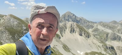 A selfie is taken by a man with weathered skin, wearing a light-colored cap and a bright yellow shirt under a blue scarf. He appears to be on a mountain, with rugged, rocky peaks stretching into the background under a clear blue sky with sparse white clouds. The landscape is arid and mountainous, with patches of green vegetation clinging to the slopes. The man's expression is neutral as he looks towards the camera. The scene suggests an outdoor excursion in a mountainous region. The city of L'Aquila is not visible in this image, and there are no other people or man-made structures depicted.