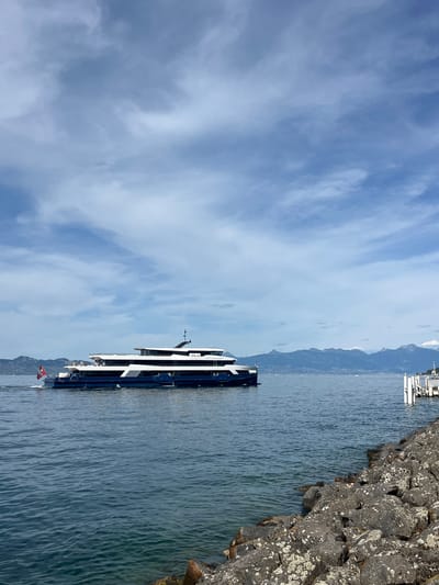 An EVIAN EXPRESS passenger ferry, painted navy blue and white, moves across a calm, blue lake under a cloudy sky. The ferry has multiple decks and is flying a Swiss flag from its stern. In the background, a range of blue-hued mountains is visible, suggesting a location near a large body of water surrounded by alpine scenery. In the foreground, a rocky shoreline slopes into the water. A white pier with wooden posts is visible to the right of the frame. The scene is likely Évian-les-Bains, France, due to the mention of the "EVIAN EXPRESS" ferry, which operates on Lake Geneva (Lac Léman), bordering France and Switzerland.