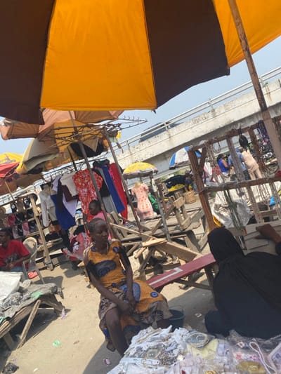 The image depicts an outdoor market scene in the city of Aiyetoto-Asogun, Nigeria, beneath an elevated concrete overpass. The foreground is dominated by a large, bi-colored market umbrella, orange and brown, partially obscuring the upper left and right.

In the immediate foreground, a young woman with dark skin and braided hair sits on a red and white wooden bench. She wears a patterned orange, blue, and black dress and faces slightly left of the camera. A black bucket rests near her feet. To her right, a person dressed in a black abaya and head covering sits on the ground with their back mostly to the viewer, positioned behind a makeshift display of various small items and jewelry, including necklaces and packaged goods, spread on white cloths.

The midground features numerous temporary market stalls constructed from rough wooden planks and poles. One prominent stall on the left displays an array of clothing, including dresses, shirts, and skirts in various colors, hanging from wooden racks. Below these garments, shoes are arranged on tiered shelves. Further back, another stall displays a light-colored dress on a mannequin-like stand. Several other individuals are visible throughout the market, some observing goods or moving between stalls.

The background is defined by a concrete overpass or bridge running horizontally across the upper portion of the frame. A light-colored vehicle is visible traveling on the overpass from left to right. The ground throughout the market appears to be a mix of packed dirt and worn concrete. The scene is illuminated by bright daylight under a clear sky, indicating an active market environment.