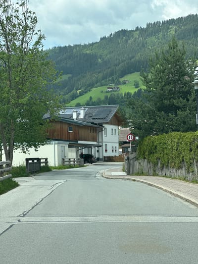 The image depicts a street scene in Reitdorf, Austria, under a cloudy sky. The foreground shows a paved road with visible white lane markings and a slight crack. To the left, a wooden fence borders a grassy area. Further into the scene, a building with a wooden facade and a white lower level is visible, featuring solar panels on its roof and a covered parking area. A dark-colored car is parked under the canopy. Adjacent to this building, another building with white walls and red-framed windows is present. A circular traffic sign with two vertical black bars on a white background within a red circle is mounted on a pole. A tall evergreen tree stands to the right of the buildings, with a dense green hedge and a wooden plank fence running alongside the road. In the background, a lush green mountain slope covered with a dense forest rises, with scattered houses visible on its lower parts.
