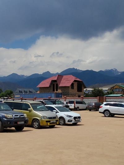 A lot of parked cars are in front of a building under construction. The building has a red roof and appears to be made of brick. In the background, there are mountains. The sky is cloudy.