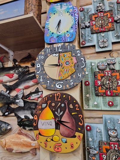 The image displays a close-up view of a collection of decorative ceramic clocks arranged on wooden shelves and a wooden wall. The clocks feature whimsical designs, with some depicting cats, owls, and other animals, as well as abstract patterns and a wine bottle motif. One clock has the word "VINO" written on it. Several ceramic fish sculptures are also visible, hanging on the wooden wall. The overall setting appears to be a shop or market stall, likely selling handmade crafts. The background consists of wooden planks and shelves, with a hint of a blue sky visible on the top left. There are no people present in the image.