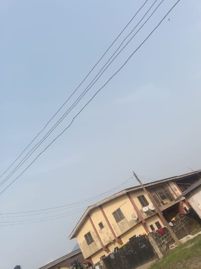 The scene depicts an urban exterior in Okere-Urhobo, Nigeria, viewed from a low, upward-tilted angle. The upper two-thirds of the image show a clear, light blue sky traversed diagonally by four parallel dark utility wires from the mid-left to the upper-right. A single, small dark bird is visible in flight in the upper-right quadrant of the sky.

The lower third of the image features a two-story residential building, predominantly beige with some darker staining and a dark brown roof overhang. The building has multiple rectangular windows with dark frames, and two white satellite dishes are mounted on its upper-story facade. A wooden utility pole supporting additional wires is positioned in the foreground on the right, partially obscuring the building. In front of the building, a low, dark, rough-textured concrete block wall or fence runs horizontally, over which various garments, including red and yellow clothing, are draped. To the immediate left of the main building, a darker, partially visible structure is present. The ground in the bottom-right corner shows patches of green vegetation. No people are visible.