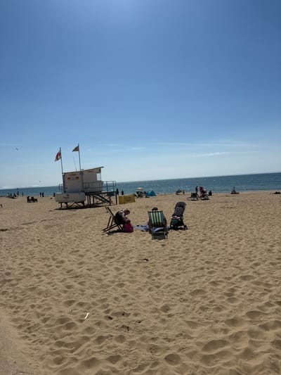 This is a vertical, eye-level shot of a sandy beach with the ocean in the background. The sky is clear and blue, suggesting a sunny day. The time of day appears to be midday or early afternoon, given the bright sunlight casting shadows. The location is Bournemouth, UK, indicated by the presence of a lifeguard tower with text that reads "Lifeguards" and the Union Jack flag.
The beach is populated with several people. In the foreground, a person is relaxing in a beach chair with a yellow and blue striped umbrella behind them. Further back, there are several other individuals and groups scattered across the sand, some sitting in chairs, others standing. A stroller is parked on the sand. The lifeguard tower is a prominent feature, situated on the beach near the water's edge. Two flags are attached to the tower. The sand is textured with footprints and ripples, evidence of recent activity. The ocean is calm and reflects the sunlight. The overall atmosphere seems relaxed and leisurely, characteristic of a typical beach day.