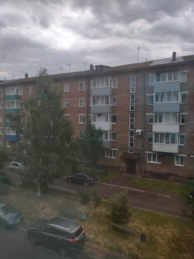 The image depicts an overcast day in a residential area, likely Osinniki, Russia. A multi-story brick apartment building with numerous windows and balconies dominates the scene. Several cars are parked on the street and in a courtyard area with patchy grass and trees. The building's facade is a mix of reddish-brown brick and white and gray balcony enclosures. Various antennas are visible on the roof of the building. In the foreground, a dark-colored SUV and a dark sedan are parked. A light-colored SUV is visible further back in the courtyard. A wooden bench and a trash receptacle are situated in the grassy area. The sky is filled with dense, gray clouds, indicating a potentially rainy or stormy day.