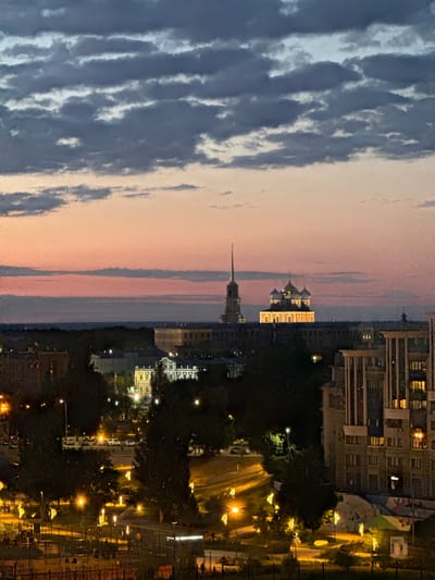 The image depicts a twilight cityscape, likely Ryazan, Russia, characterized by a dramatic sky and illuminated architectural landmarks. Dark, textured clouds occupy the upper portion of the frame, transitioning into a soft gradient of peach and pink hues near the horizon. Below the sky, a prominent spire pierces the dusk, suggesting a significant religious or historical structure. To its right, a domed building, possibly a cathedral or church, is brightly lit, its golden domes glowing against the darkening sky.
The middle ground reveals a dense arrangement of buildings, some with illuminated windows, interspersed with dark trees. The illumination from streetlights and interior building lights creates a warm glow across the urban landscape, highlighting streets and pathways. In the foreground, the scene transitions to a lower level of the city, with more intensely lit trees, street lamps, and building facades. A large, modern apartment building with multiple windows is visible on the far right, with some windows lit. The overall impression is one of a city coming alive with artificial light as natural light fades. There are no discernible people in the image.