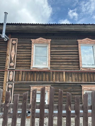 This is a medium shot, low-angle, exterior photograph of a wooden house with decorative window frames and a corrugated metal roof. The house has horizontally oriented wooden siding. There are two windows visible on the upper story, each with ornate pink trim. A vertical wooden decorative element with carved circular and diamond motifs is attached to the left side of the house. A grey pipe extends from the left side of the roof. A dark brown wooden fence made of vertical planks is in the foreground. The sky above is cloudy with patches of blue. The scene appears to be in Nogornyi, Russia. There are no people or animals in the image.