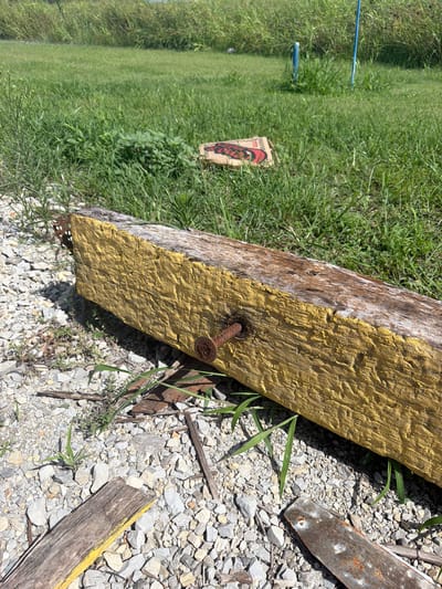 The image depicts an outdoor scene on a sunny day in Cardwell, United States. In the foreground, a weathered, rectangular wooden beam lies on a bed of gravel and scattered debris, including broken pieces of wood and a rusty metal strip. The beam is partially painted yellow, with the paint peeling in places. A single, large rusty nail protrudes from the side of the beam. Behind the beam, a grassy field stretches into the background, with tall weeds and some sparse green foliage. In the mid-ground, a flat, rectangular object with red and black markings lies on the grass. Further in the background, two blue posts are visible amidst the tall grass. The overall environment suggests an unkempt, rural, or semi-rural setting. There are no people or animals visible in the image.