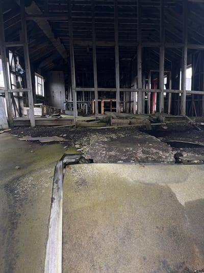 The image displays the interior of a dilapidated wooden structure. The framework of the building is visible, with beams and supports creating a grid-like pattern. Light streams through gaps in the structure, highlighting the decay. Concrete and debris fill part of the frame. The overall impression is one of abandonment and disrepair.