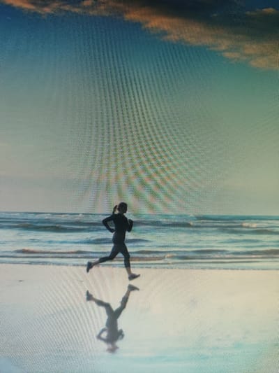 A silhouetted figure runs along a wet beach, their reflection clearly visible on the smooth, reflective surface. The figure is dressed in athletic wear and appears to be a woman. The ocean stretches out behind her, with gentle waves lapping at the shore. The sky above is a gradient of blue, with streaks of orange and pink clouds near the horizon, suggesting either sunrise or sunset. The overall atmosphere is serene and tranquil, evoking a sense of solitude and peacefulness. The location is identified as a beach in Tursunzoda, Tajikistan.