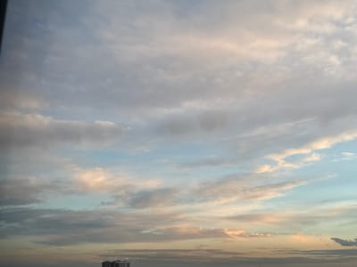 The image shows a cloudy sky with soft pastel colors, suggesting either sunrise or sunset. The clouds are varied in texture and density, with some appearing wispy and others more solid. Patches of light blue sky are visible between the clouds. In the lower portion of the image, a cityscape with several buildings is discernible, including a tall, multi-story structure that appears to be an apartment building. The scene is calm and serene, with no visible people or animals. The overall impression is one of a peaceful moment in an urban environment as day transitions to night, or vice versa. The specific location in Бордуки, Russia, cannot be determined from the visual information alone.