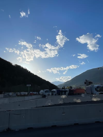 The image depicts a row of trucks parked along a road in Freienfeld - Campo di Trens, Italy. The trucks are of varying colors and sizes. The road is bordered by a concrete barrier on one side. On the other side of the trucks is a hillside covered with trees. Above the hillside is a blue sky with scattered white clouds. A contrail from an aircraft is visible in the sky. The sun is shining brightly.