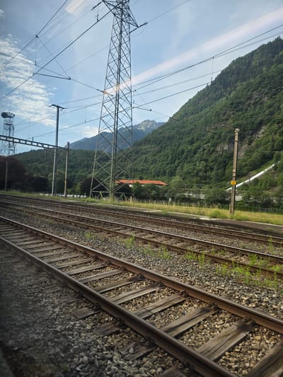 The image shows a view of multiple train tracks in Lavorgo, Switzerland, receding into the distance. The tracks are laid on a bed of gravel, with sparse green vegetation growing between the sleepers. Beyond the tracks, a steep, densely forested mountain slope rises dramatically. The sky is partly cloudy with a clear blue sky visible. Several tall electricity pylons and overhead power lines crisscross the upper part of the image, indicating the presence of significant electrical infrastructure. A building with a red roof is partially visible nestled at the base of the mountain. A bright white pipe or channel runs down a section of the mountain to the right, possibly for water management. The scene is captured from the perspective of a moving train, as evidenced by the slight blur and the angle of the tracks. There are no people visible in the frame.