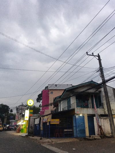 The image depicts a street scene in Phước Hòa, Vietnam, under an overcast, grey sky. Numerous black utility lines crisscross the upper frame, extending from a concrete utility pole positioned on the right side. The foreground features an asphalt street with a partial white painted line. On the left, a series of low-rise commercial structures display illuminated signs, notably a prominent circular yellow sign featuring a dark, panda-like figure, and a vertical yellow and green sign displaying "24H". A yellow vehicle and several motorcycles are partially visible further down the street on the far left, with indistinct figures mounted on them. Adjacent to the illuminated businesses, a two-story building with a light-colored upper facade and a pitched, green-tiled roof presents a ground-floor commercial space with blue-painted details and visible red plastic chairs, identified by a banner that reads "NASI GORENG AYAM". A red and white striped flag is affixed vertically to the right side of this building. In the background, a multi-story building exhibits distinct pink and pale orange sections, featuring small, square windows. Trees are visible behind the buildings. No direct human actions or interactions are clearly discernible in the immediate foreground.