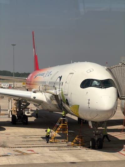 This image, captured from inside an airport terminal in Jianyang City, China, depicts a large passenger aircraft, likely a Boeing 787 Dreamliner due to its distinctive cockpit windows, parked at a gate. The aircraft is connected to a jet bridge, indicating preparation for boarding or recent arrival.

Several ground crew members, wearing high-visibility vests, are actively working around the plane. One person is visible near the nose landing gear, while others are situated near the main landing gear and along the fuselage, performing checks or handling ground support equipment like yellow mobile stairs.

The aircraft features a colorful livery, transitioning from shades of red and orange on the tail and rear fuselage to lighter tones with stylized designs towards the front. The tail fin is prominently red. The scene takes place during a clear and bright day, evidenced by strong shadows cast by the aircraft and equipment on the tarmac.

The concrete apron is marked with various parking position indicators for different aircraft types, including "A380," "B747," "A340," "B777," "A330," and "B787," along with a large yellow 'X' and other directional markings. A yellow sign displaying "181" is visible near the aircraft's wing. The photo provides a candid view of typical airport ground operations.