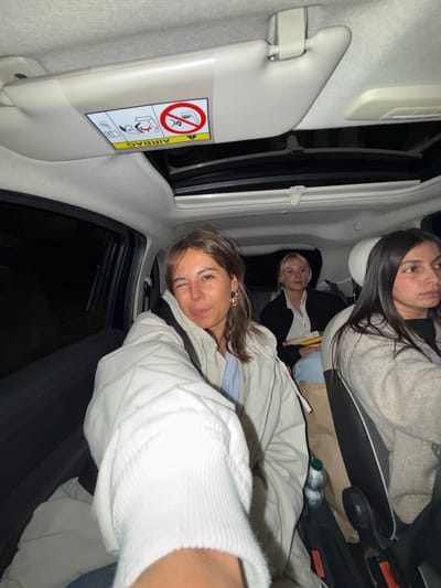 Inside a car, three women pose for a photo. The first woman, positioned in the front seat, smiles with a beige jacket. Behind her, another woman with dark hair wears a gray sweater. A third woman with blonde hair is visible in the back seat, between the two. A sun visor with warning labels is visible on the side of the car. Water bottles and other items are on the seat.