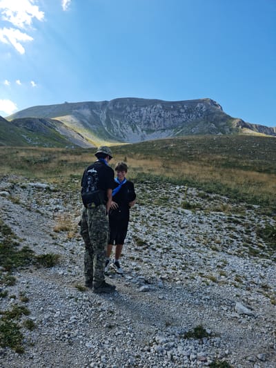 In the city of L'Aquila, Italy, two individuals are standing on a gravel path in a mountainous terrain. The person on the left is wearing camouflage pants, a black t-shirt with a graphic print, a hat, and a black fanny pack. The person on the right is wearing shorts, a black t-shirt, and sneakers. Both individuals are wearing blue bandanas around their necks. They are facing each other and appear to be conversing. In the background, there is a large, rocky mountain with sparse vegetation under a clear blue sky with some clouds. The sun is casting light and shadows on the landscape. The environment is natural and appears to be a hiking trail.