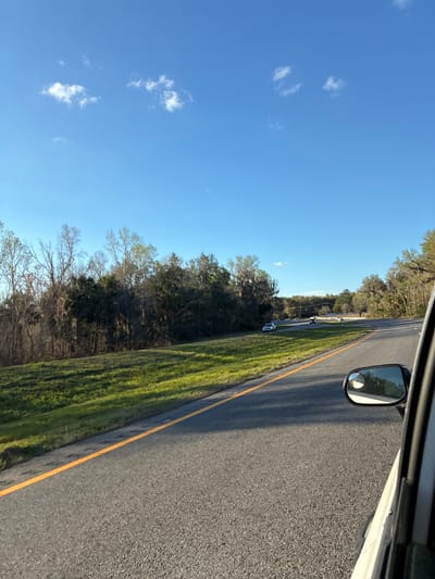 The image depicts a daytime scene on a multi-lane asphalt highway, likely in the city of Micanopy, United States. The perspective is from inside a moving vehicle, with the right side mirror and a portion of the white vehicle's window frame visible in the lower right. The sky is bright blue with scattered small, white cumulus clouds.

The highway features at least two lanes traveling in the same direction, marked by a continuous solid yellow line on the left side of the visible roadway. The asphalt surface is dark gray. To the left of the highway, a grassy embankment slopes upwards towards a dense line of diverse trees, which include both bare deciduous trees and evergreens, some adorned with Spanish moss. The terrain is gently undulating.

Several vehicles are observed on the highway ahead, all appearing to be in motion. A light-colored sedan is visible in the left lane in the midground, followed by at least two other vehicles further in the distance as the road curves slightly to the right. Sunlight creates distinct shadows on the road surface, suggesting a clear day. No individuals are distinctly visible.