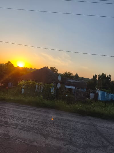 The image shows a rural landscape at sunset, possibly in or near Paladea, Moldova. In the foreground, a cracked asphalt road runs diagonally across the frame. To the left of the road, tall green grass and foliage line the embankment. In the mid-ground, a cluster of old, weathered houses with dark, sloped roofs are visible. One house has green siding and is partially covered in vines. Satellite dishes are mounted on the roofs of some structures. Trees and other vegetation are scattered around the houses and in the background. The sky is a gradient of warm colors, transitioning from a bright orange and yellow at the horizon where the sun is setting, to a pale blue overhead. Electrical wires are strung across the upper portion of the image. The scene evokes a sense of tranquility and the end of a day in a rural setting.