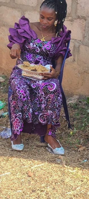 An adult female is seated outdoors on a blue plastic chair in Alayi, Nigeria. She wears a floor-length, vibrant purple and black patterned gown with ruffled sleeves. Her hair is styled in dreadlocks or braids, adorned with gold earrings and a gold necklace featuring a distinct pendant. She is holding a silver-colored rectangular serving platter containing a meal composed of light-brown granular food, possibly rice, mixed with darker components. Her right hand, holding a spoon or similar utensil, is positioned over the food as if actively eating. Her gaze is directed downwards towards the platter. She is wearing light-colored, possibly silver, flat shoes with bows. The ground beneath her is dry earth with sparse, light-brown vegetation and scattered debris. Behind her is an unpainted wall constructed from light-brown or beige bricks or cinder blocks.