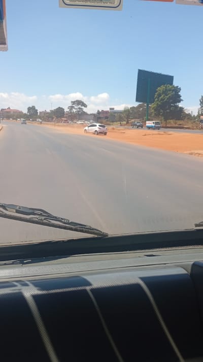 A dusty road stretches ahead under a clear blue sky. A white car kicks up dust on the road shoulder. Several other vehicles are visible in the distance. A large, blank billboard rises alongside the road, adjacent to a tree. The view is from inside a vehicle, as indicated by the windshield wipers and part of the dashboard.