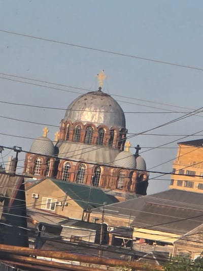 The image shows an exterior view of a large, ornate domed building, likely a church, with several smaller domes. The architecture is detailed, with arched windows and brickwork. Several electrical wires crisscross the foreground and midground, obscuring parts of the structure. In the background, there is a tall, yellow-orange building and other urban structures, suggesting a city setting. The sky is clear and blue. The scene is likely Khasavyurt, Russia, given the architectural style commonly found in Russian Orthodox churches and the presence of overhead wires typical of older urban infrastructure. There are no people visible in the image.