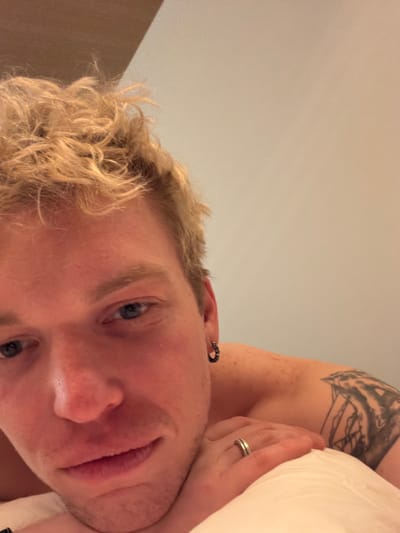 The image displays a close-up of a male individual, observed in an indoor setting in Kutchan, Japan. The subject possesses light-colored, curly hair and blue eyes. He is positioned, likely lying down, with his head and upper torso visible. A dark-colored hoop earring is present in his right ear. On his left shoulder, a tattoo depicting a sailing ship or similar nautical vessel is visible. A silver-colored band ring is worn on the ring finger of a hand, resting on a white pillow in the lower-left foreground. The background consists of a plain, light-colored wall.