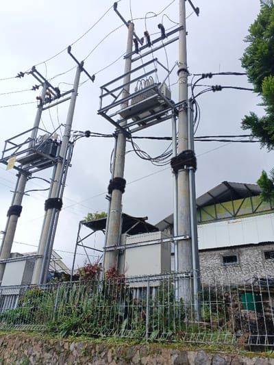 The scene in Uato Carabar, Timor-Leste, depicts an electrical utility infrastructure under an overcast sky. Three concrete utility poles rise vertically, supporting a network of power distribution equipment. Two prominent electrical transformers are mounted on galvanized steel platforms attached to the poles; the transformer on the left is darker, while the central transformer is lighter grey with visible cooling fins. Numerous black electrical power lines and communication cables are strung between the poles, connected via insulators and cross-arms. Several sections of the poles and cable bundles are wrapped with black, coarse, fibrous material. At ground level, a galvanized wire mesh fence runs horizontally, positioned above a dry-stacked stone retaining wall. Behind the fence, green shrubs and reddish plants are visible. In the background, sections of buildings include a structure with a white facade and a grey corrugated metal roof supported by green trusses, and a rough-hewn stone building featuring dark, recessed window openings. No individuals or discernible actions are present within the frame.