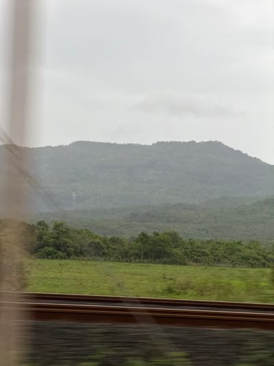 The image captures a misty, overcast day with a prominent mountain range in the background. The mountains are covered in lush green vegetation, and a thin waterfall is visible cascading down the mountainside on the left. In the middle ground, there is a clear demarcation between a grassy field and a dense forest. A wire fence runs along the edge of the field, bordering the trees. In the foreground, a set of train tracks, appearing to be moving due to motion blur, runs horizontally across the frame. The overall scene suggests a rural or natural landscape, possibly viewed from a moving vehicle. The location is Saphale, India.
