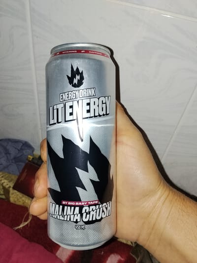 A hand is holding a silver energy drink can with a black and white design. The can has text that reads "ENERGY DRINK," "LIT ENERGY," "MALINA CRUSH," and "450 ML." The background appears to be a tiled wall. The image does not provide any information about the city of Dagestanskiye Ogni, Russia, or any specific location.