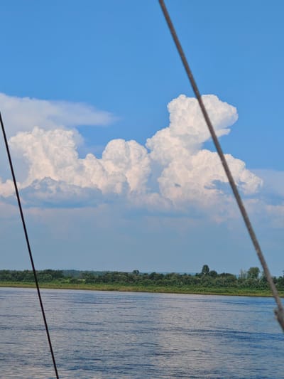 A wide, blue river flows from left to right in the foreground, with ripples on its surface. The riverbank is lined with a dense, green forest. Above the treeline, large, white cumulus clouds fill the bright blue sky. Two dark ropes are visible in the foreground, angled from the bottom left and top right corners of the image, suggesting the perspective is from a boat. The scene is a natural landscape, likely a river in a rural area.