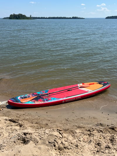 A red stand-up paddleboard with a colorful design is partially submerged in the shallow water at the edge of a sandy beach. A paddle with an orange blade rests on top of the board. The paddleboard has the text "SHAMAN CITY" and "RAVE BOARD" printed on it. The water is calm with a slight ripple. In the background, a large body of water stretches to the horizon, with a tree-covered island visible in the distance. The sky is blue with a few scattered clouds. The location is the city of Borovoye, Russia.