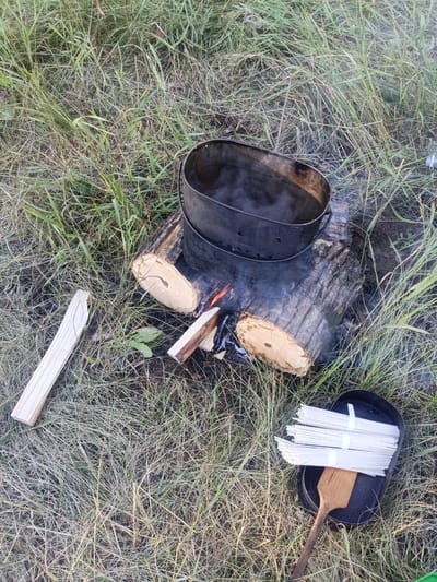 The image depicts an outdoor scene, likely in a grassy area with dry grass and some green vegetation. In the center of the frame, a black metal pot is placed on top of a log, which is part of a makeshift campfire. The pot contains steaming water, indicating it is being heated over a fire. Flames and smoke are visible beneath the pot, emanating from burning wood. To the left of the pot, a long, splintered piece of wood lies on the ground. To the right, a black metal pan holds several bundles of uncooked spaghetti, tied with white bands. A wooden spatula rests on top of the spaghetti. The overall impression is one of outdoor cooking preparation, possibly camping or a picnic. The location is stated to be in the city of Letniki, Russia.