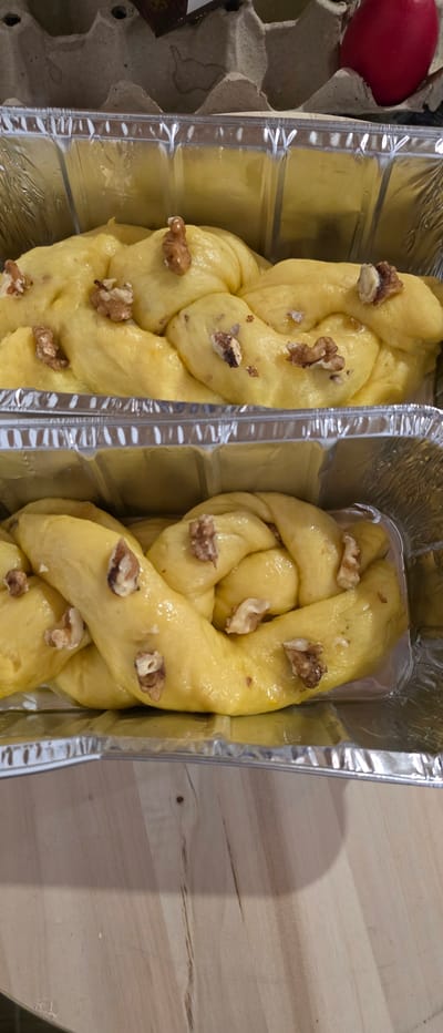 The image shows two aluminum foil containers, each containing a braided, yellow bread topped with walnuts. To the left of the containers, partially visible, is a cardboard egg carton with a single red egg. The setting appears to be a wooden surface, possibly a table. The scene is likely indoors. There is no indication of this being located in Novachene, Bulgaria.