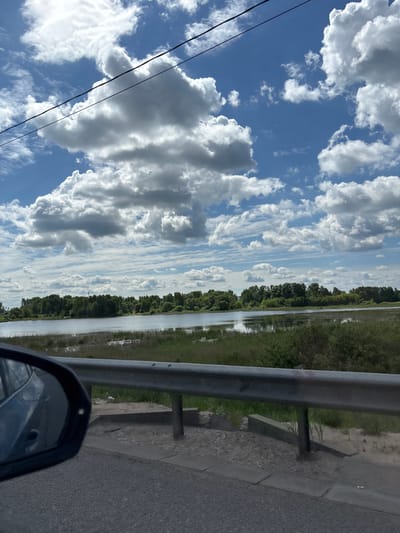 This image captures a vibrant outdoor scene from the perspective of a vehicle, likely in motion. The dominant feature is the expansive sky, showcasing numerous large, fluffy white cumulus clouds against a bright blue background. Some darker, denser clouds are also present, adding depth to the sky. Two dark power lines diagonally cross the upper left part of the frame.
Below the sky, a landscape featuring a road, guardrail, and a body of water unfolds. On the right side, an asphalt road is visible, running alongside a sturdy grey metal guardrail supported by concrete posts. Beyond the guardrail, green grass and other vegetation line the edge of a calm body of water, possibly a river or canal, which stretches horizontally across the midground. The water beautifully reflects the sky and clouds, creating a striking mirror effect. A dense line of dark green trees flanks the far bank of the water body, extending into the distance.
In the bottom right corner, a portion of a car's side-view mirror is visible, reflecting elements of the vehicle and its surroundings. A small, white bird is perched on the ground near the guardrail, situated between it and the water.
The scene depicts bright daylight, indicative of a clear and sunny day with scattered clouds. The overall impression is one of peaceful transit through a scenic, likely rural or semi-rural, area, consistent with the location context of Staroye Melkovo, Russia. No visible text is present in the image.