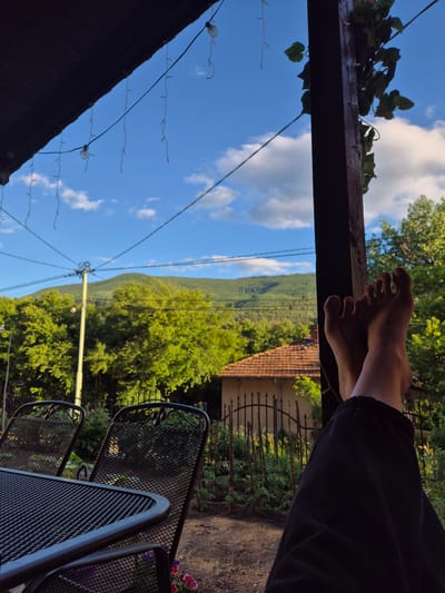 The image depicts a perspective from a covered outdoor seating area, likely a porch or patio, in Iskrets, Bulgaria. In the foreground, a person's bare feet are propped up on a dark-colored wooden post, wearing black sweatpants. The feet are illuminated by natural light, highlighting the skin tone and toenails. Two dark, metal mesh chairs and a matching table are visible to the left, suggesting a dining or relaxation setup.
The background reveals a rural landscape. Lush green trees dominate the mid-ground, with a traditional house featuring a terracotta tiled roof and a rustic fence visible behind the chairs. Beyond the trees, a gentle, green mountain slopes upward against a bright blue sky scattered with white clouds. String lights, some illuminated and some not, hang from the covered area, adding a decorative element. The overall impression is one of a peaceful, outdoor setting, possibly during the late afternoon or early evening, given the quality of light and shadow.