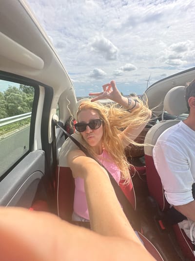 A blonde woman with sunglasses is taking a selfie in a convertible car. She is wearing a pink top and has her hair blowing in the wind. Her left hand is raised, making a peace sign. A person wearing a white shirt is visible in the passenger seat. The car is on a road with trees and a wind turbine in the background. The sky is cloudy. The image is taken in Nouvion-le-Comte, France.