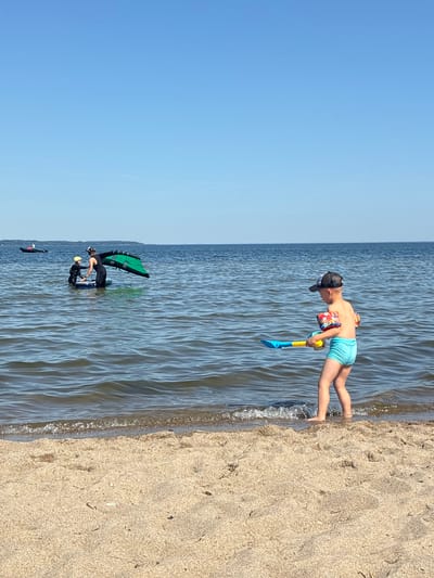 The image depicts a sunny day at the beach, likely in Rewa, Poland. The scene is divided between a sandy shoreline and the calm, blue water. In the foreground, a young boy wearing swim trunks and floaties is standing in the water, holding a toy shovel. His posture suggests he is about to play or has just finished playing. Further out in the water, two individuals are engaged in a water sport, possibly windsurfing or wing foiling. One person is kneeling on a board with a large green wing, while another person appears to be assisting them. The water is slightly choppy, indicating a gentle breeze. The sky is clear and blue, suggesting good weather conditions for outdoor activities. The sand is light brown and appears dry, with some footprints visible. The overall impression is one of a pleasant summer day spent enjoying recreational activities by the sea. No visible text is present in the image.
