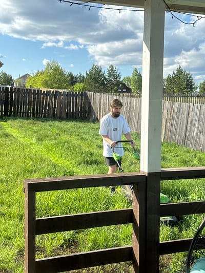 This image depicts an outdoor scene in a backyard, likely in Kuda, Russia, captured during bright daylight under a partly cloudy sky. The main subject is a man with a beard, wearing a white t-shirt with a "P.A.M." logo on the left chest, and dark shorts. He is actively engaged in trimming grass using a bright green and black electric string trimmer.
The man is positioned beneath a wooden structure, possibly a porch or deck, which features prominently in the foreground and frames the scene. A white wooden support post is visible on the left, adorned with string lights. Dark brown wooden railings and framing of the structure are seen on the right and across the bottom of the image. The ground is covered in lush green grass, suggesting a well-maintained or recently overgrown lawn. In the background, a rustic wooden fence stretches across the width of the yard, with various trees and a glimpse of a building visible beyond it. The clear shadows indicate a sunny day, providing strong contrast to the bright green grass and blue sky. The man appears focused on his task.