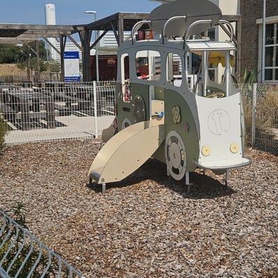 This image depicts an outdoor playground in La Palme, France, with a whimsical play structure resembling a vintage camper van. The van is primarily green and white, featuring a curved slide, a steering wheel, and decorative wheels and flowers. The ground is covered with wood chips, typical for playground safety. In the background, a white metal fence encloses picnic tables and wooden seating areas, suggesting a recreational or dining space. Beyond the fence, a large white industrial silo labeled "ENGIE" is visible, indicating the presence of an industrial facility in the vicinity. The sky is clear and blue. There are no people visible in the immediate foreground or on the play structure. The overall scene is one of a well-maintained outdoor public space.