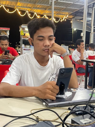 This image depicts an evening scene in Lhok Mereubo, Indonesia, within an illuminated, semi-open-air dining area.

In the foreground, a young adult male with dark hair, wearing a white t-shirt and a silver wristwatch, is seated at a light-colored table. He has a dark earbud in his left ear. His left arm rests on the table, with his left hand supporting his chin. His right hand holds a dark-colored smartphone, which is propped upright by a black binder clip on a closed silver-grey laptop. A white USB flash drive is inserted into the right side of the laptop. Numerous black and green cables are loosely arranged across the table in front of him.

To the left, a second male in a red t-shirt is seated, focused on a black smartphone he holds. Behind him, a metal-framed food stall or cart with shelves displaying items and a yellow bag is visible, with a motorcycle partially obscured behind it. To the right, a third male in a white t-shirt with a "LFP" logo is seated at a separate table, resting his chin on his right hand and looking towards the right. Other indistinct figures and a vehicle are visible in the dim background. The area is illuminated by overhead string lights with exposed bulbs, creating a warm glow against the dark night sky.