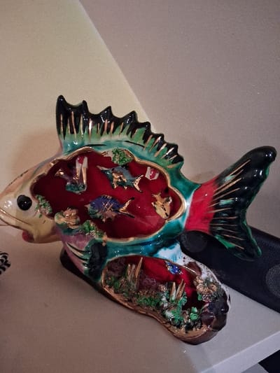 A colorful fish-shaped decorative piece with an open interior. The fish has black and green fins with gold accents, a red head and tail, and a white bottom. Inside the open fish body, there are miniature landscapes with trees, plants, and possibly other fish. The interior is primarily red and appears to be velvet-lined. It's positioned against a plain beige surface.