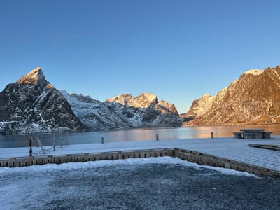 This image depicts a winter scene in Hamnøya, Norway. The foreground features a snow-dusted wooden pier extending across the lower portion of the frame, turning right into a wider platform. On the left section of the pier, three wooden posts with taut chains strung between them are visible. On the wider platform to the right, a stone picnic table with attached benches is present, covered in a thin layer of snow. The ground immediately in front of the pier exhibits patches of dark ice and snow. A calm body of water, likely a fjord, occupies the midground, reflecting a clear blue sky and warm golden light. In the background, majestic, rugged mountains, heavily snow-capped, rise steeply from the water's edge. The peaks on the left are partially illuminated by golden sunlight, while the mountains towards the right are bathed in a prominent warm, golden glow from a low-angle sun. The sky above is clear and light blue. No individuals are visible, and no discernible actions or interactions are occurring. The environment is characterized by clear, cold, winter conditions.