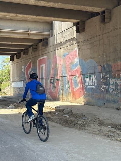 In Barranquilla, Colombia, under a concrete overpass, a person rides a bicycle on a paved road. The overpass is supported by thick concrete columns and beams, with visible rebar and concrete construction details. The wall beneath the overpass is extensively covered in graffiti, featuring large, stylized letters in red, blue, and white, with some areas of faded or partially obscured tags.
The cyclist, seen from behind, wears a blue long-sleeved shirt, dark pants, and a black helmet. A small, dark backpack with a white logo that reads "SURA" is slung over their shoulders. The cyclist is on a dark-colored bicycle with silver handlebars and black tires. Their right foot is on a pedal.
The road surface is asphalt, with a sandy, uneven area to the right of the cyclist, littered with small rocks and debris. The environment appears somewhat unkempt, with a general sense of urban infrastructure and decay. Sunlight casts shadows from the overpass onto the wall and the road, indicating it is daytime. Trees with green foliage are visible in the background on the left, suggesting a vegetated area beyond the overpass.
