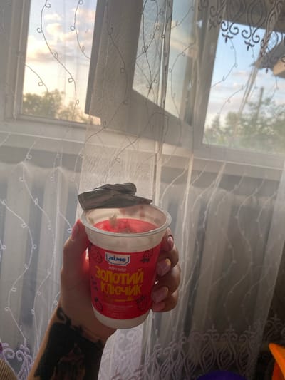 A hand holds a small, open yogurt cup with a red label that reads "ЗОЛОТИЙ КЛЮЧИК" in yellow Cyrillic letters. The yogurt appears pink and creamy with visible pieces. The background consists of a window with sheer white curtains patterned with dark, looping designs. Outside the window, a soft, cloudy sky with hints of sunset colors is visible, along with the silhouettes of trees. The scene appears to be indoors, likely a domestic setting, in the city of Ihnatpil, Ukraine.