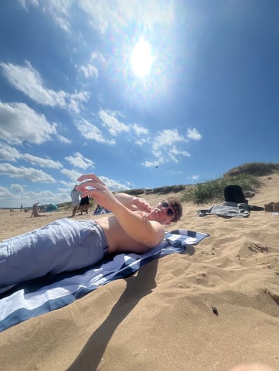 A man lies on a sandy beach beneath a sunny sky dotted with clouds. He wears sunglasses and light-colored pants, with a blue and white striped towel draped beside him. His bare torso is visible. In the background, other beachgoers and discarded clothing are scattered on the sand near the edge of a grassy dune. The camera angle is low, looking upwards towards the subject and sky.